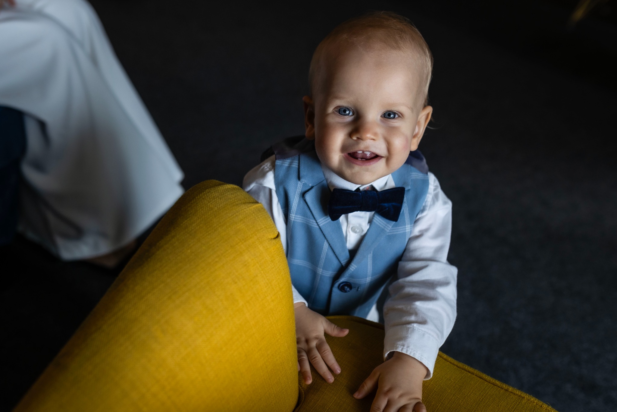 Uśmiechnięty chłopiec w niebieskiej kamizelce i muszce, oparty o żółty fotel, patrzy w obiektyw.