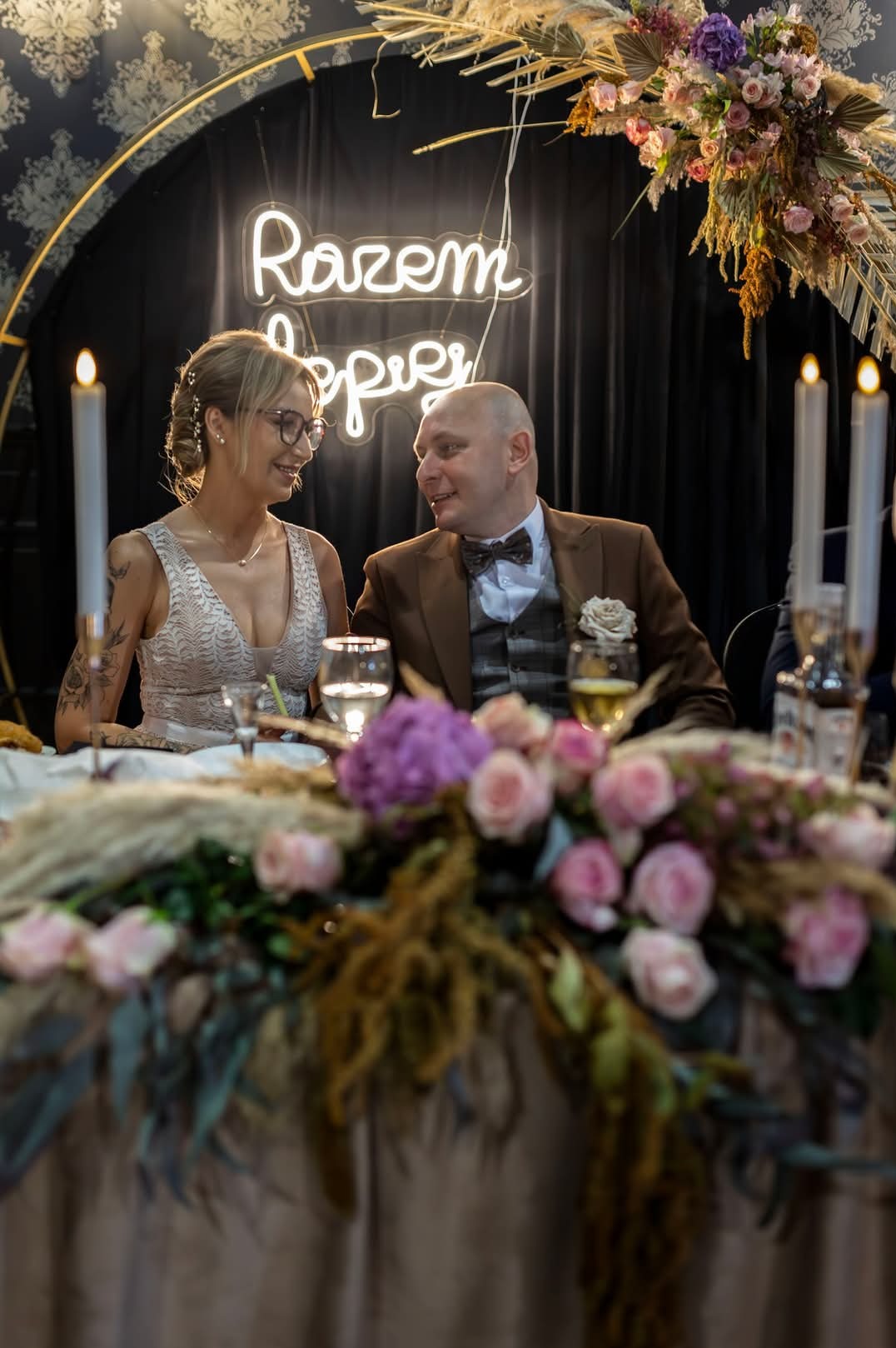 Para młoda siedząca przy bogato zdobionym stole weselnym w Piotrkowie Trybunalskim, w tle neonowy napis 'Razem Lepiej' i świece.