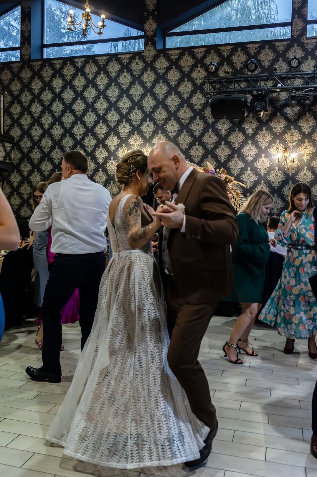 Para młoda tańczy pierwszy taniec na weselu, panna młoda ma tatuaż na ramieniu i suknię z koronkowym wzorem, pan młody w brązowym garniturze.
