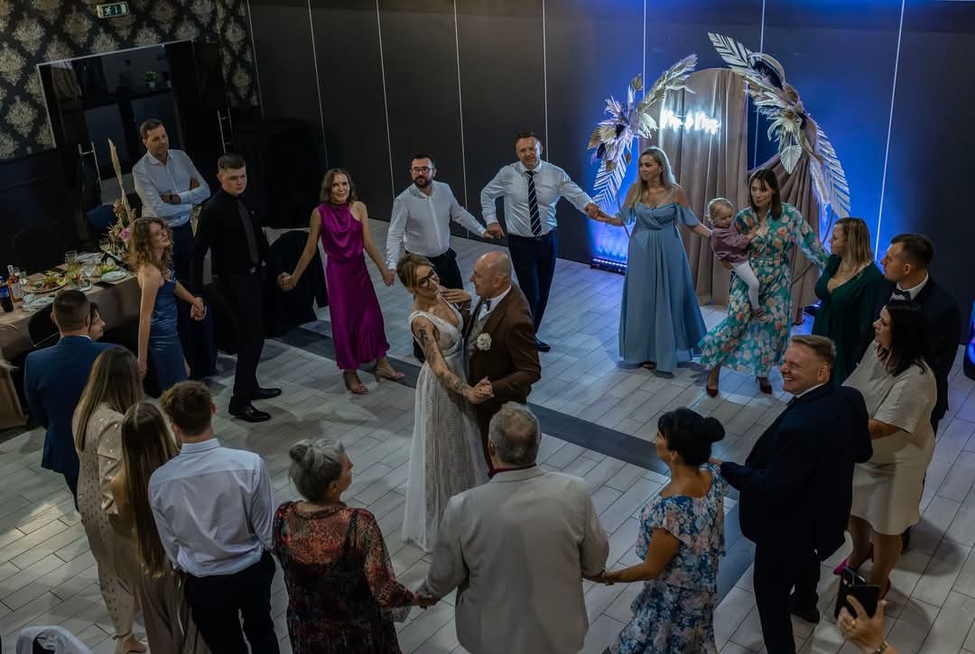 Para młoda tańczy otoczona gośćmi w trakcie wesela, dekoracja z piór i neonowe światło w tle.