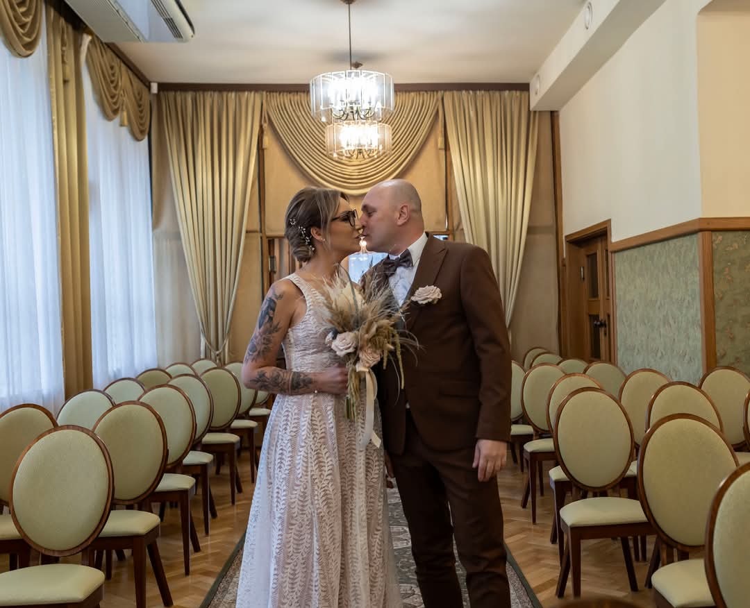 Pocałunek pary młodej w sali ślubów z rzędami krzeseł i dekoracyjnymi zasłonami w tle, panna młoda z tatuażami na ramionach trzyma bukiet