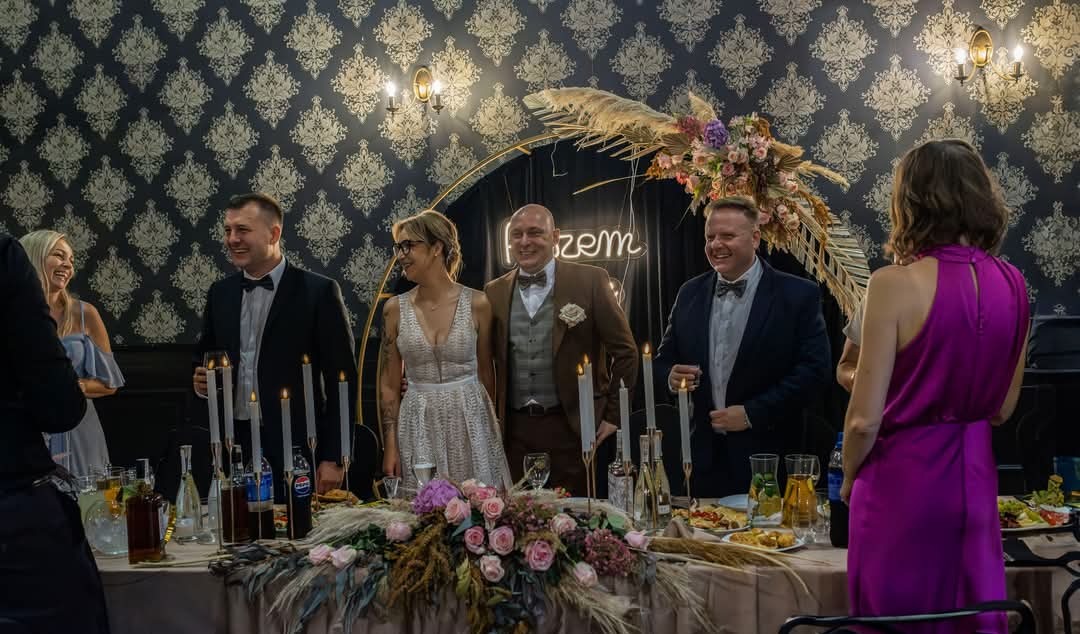 Para młoda uśmiechnięta za stołem weselnym z dekoracjami kwiatowymi i świecami, goście w tle, dekoracyjne tło ze wzorem.