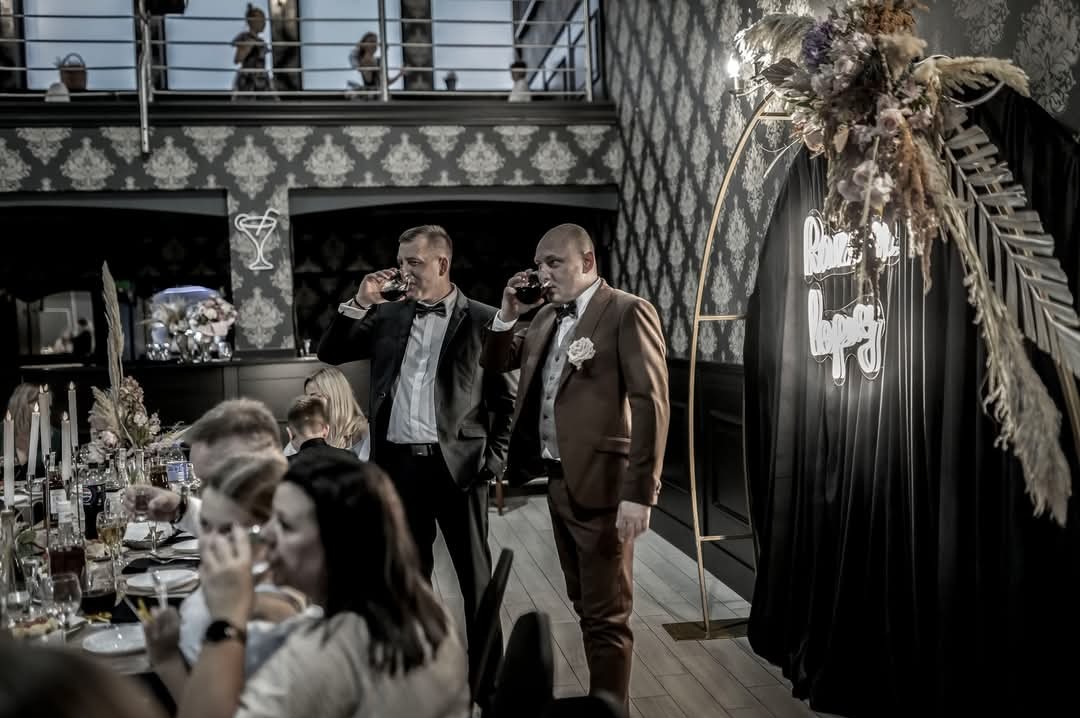 Dwaj mężczyźni w garniturach piją wino podczas uroczystości, w tle dekoracja z kwiatów i czarnej tkaniny z neonowym napisem, widoczny stół z zastawą i świecami.