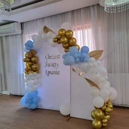 Odlotove&Balonove - Dekoracja balonowa w kolorach białym, złotym i błękitnym, w kształcie łuku, umieszczona na białym tle z napisem 'Chrzest Święty Ignasia', w sali z parkietem i zasłonami.