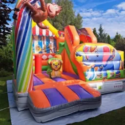 Kolorowy dmuchany zamek z motywami zwierzęcymi: małpa na tęczowym łuku, lew na platformie i słoń na zjeżdżalni, rozłożony na trawie na białej plandece.