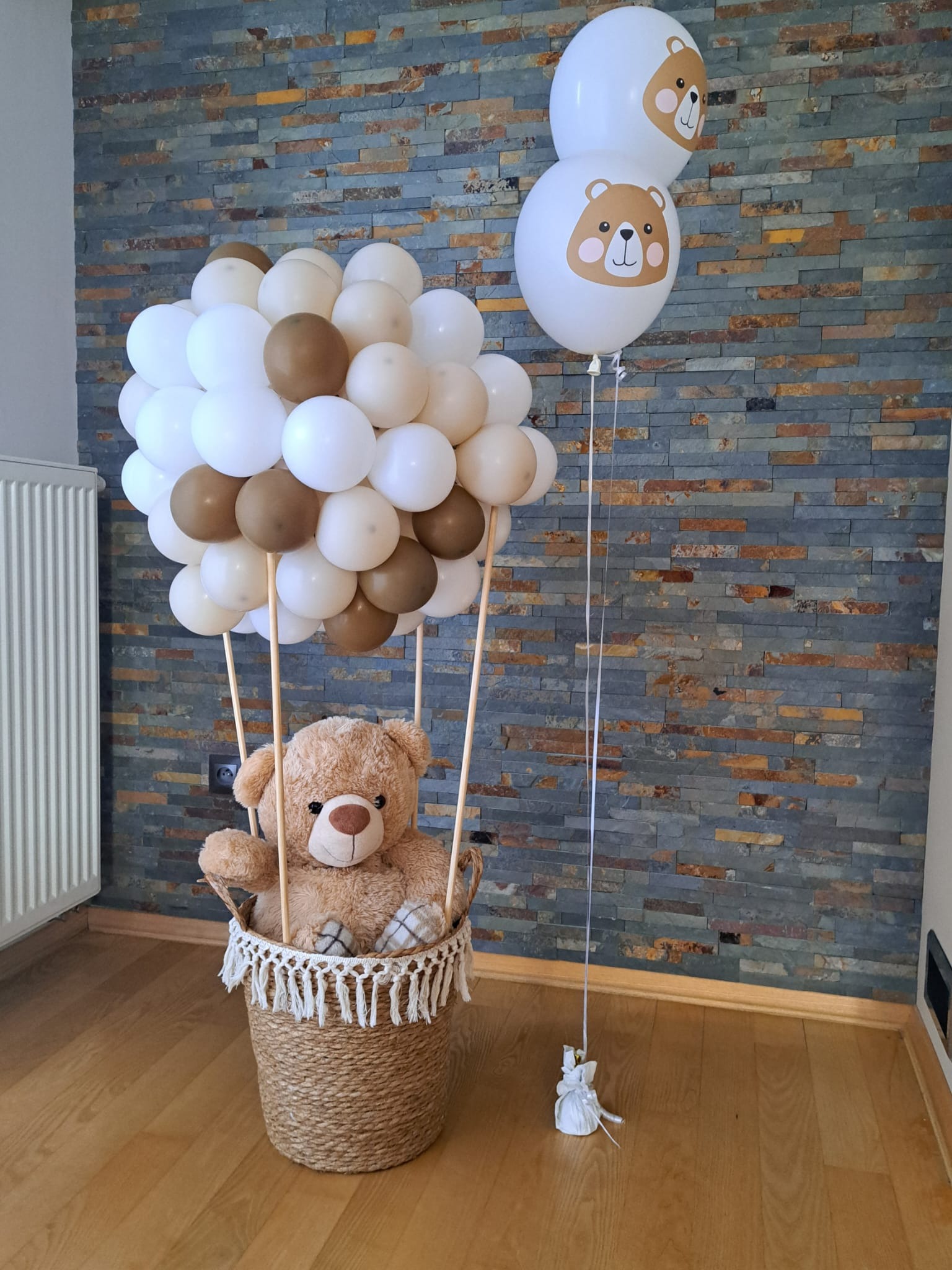 Kompozycja balonowa z pluszowym misiem w koszu imitująca balon na ogrzane powietrze, uzupełniona dwoma balonami z nadrukiem misia, na tle ściany z kamienia dekoracyjnego.