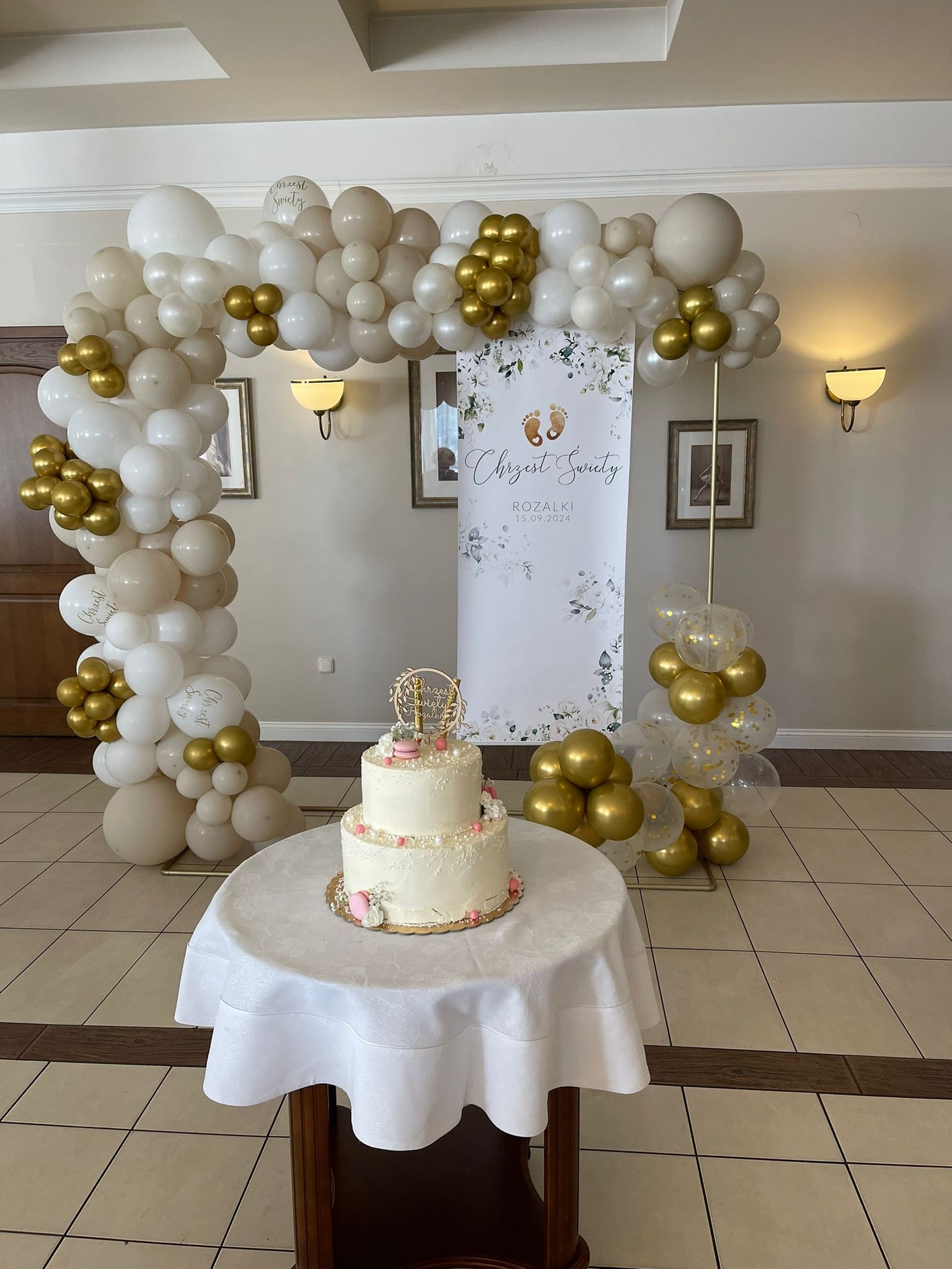 Dwupoziomowy tort z białym kremem i różowymi akcentami na stole przykrytym białym obrusem, w tle dekoracja z białych, beżowych i złotych balonów tworzących łuk oraz baner z napisem 'Chrzest Święty...