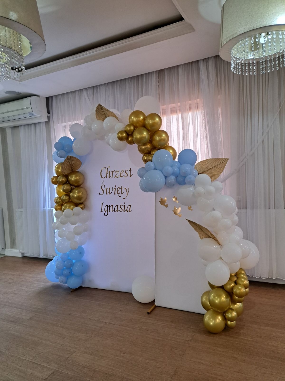 Dekoracja balonowa w kolorach białym, złotym i błękitnym, w kształcie łuku, umieszczona na białym tle z napisem 'Chrzest Święty Ignasia', w sali z parkietem i zasłonami.