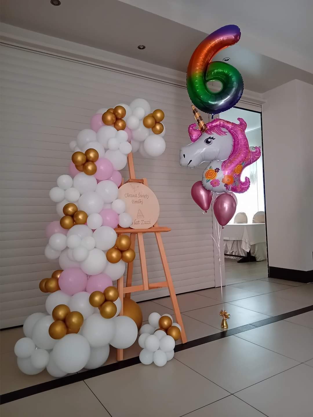 Dekoracja balonowa na Chrzest Święty Emilii: biało-różowo-złota girlanda balonowa oparta o drewniany stojak z napisem, obok balony foliowe w kształcie jednorożca i cyfry 6 w kolorach tęczy.