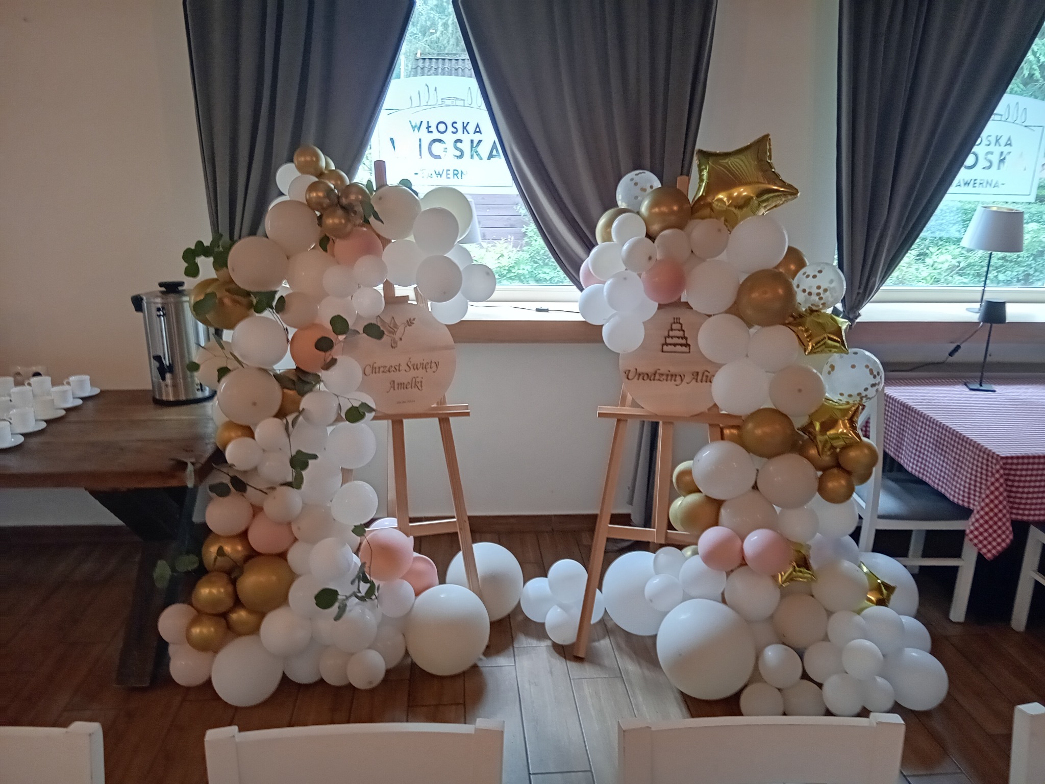 Dekoracja z białych, złotych i różowych balonów na drewnianych stojakach, z napisem 'Chrzest Święty Amelki' i 'Urodziny Alicji', ustawiona w restauracji z widokiem na okno.
