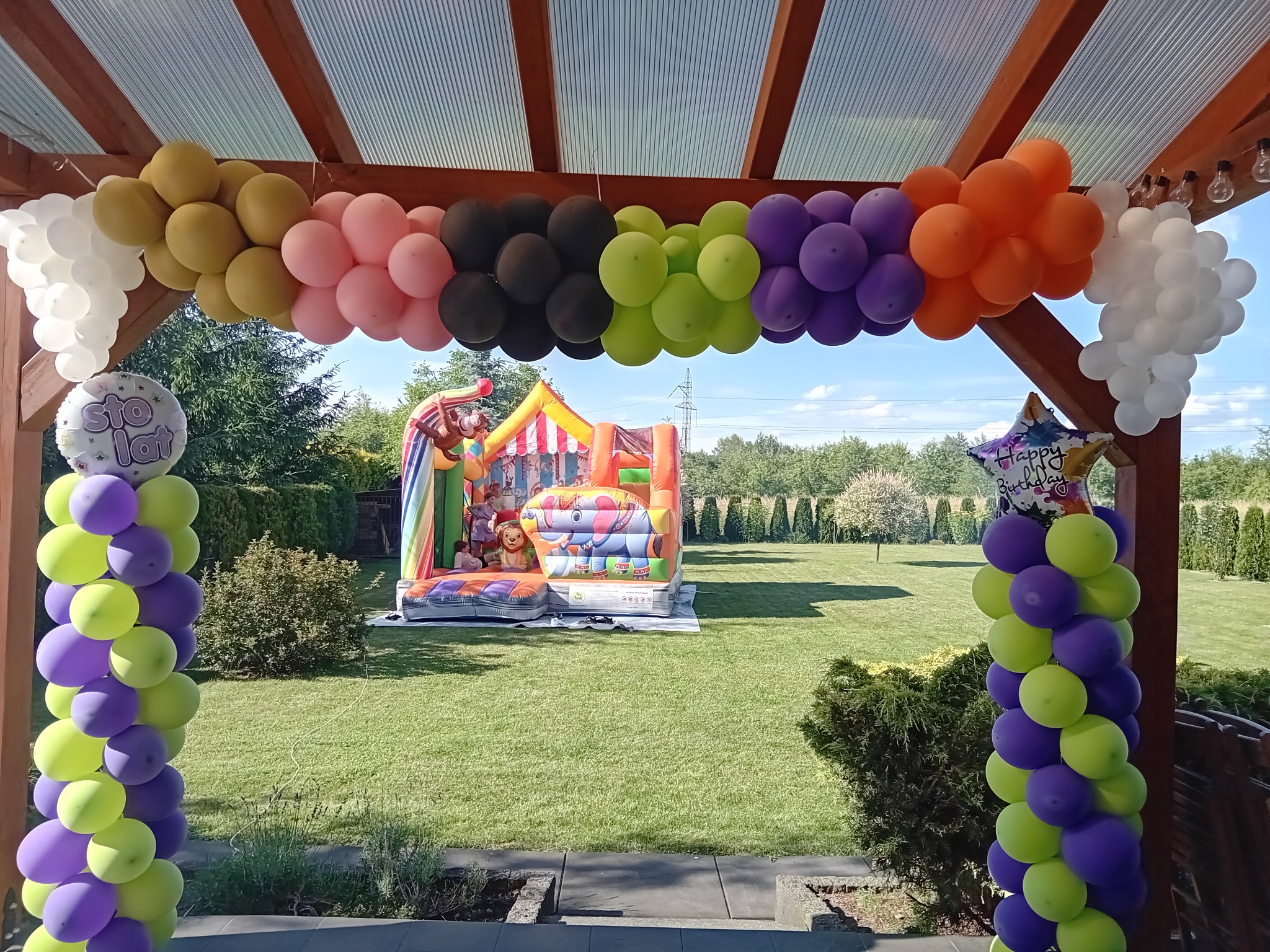 Kolorowy dmuchany zamek ze słoniem i małpą, ustawiony na trawniku, widoczny przez dekoracyjną bramę z balonów w różnych kolorach. Na pierwszym planie balony z napisem 'Sto lat' i 'Happy Birthday'.