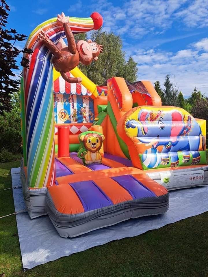Kolorowy dmuchany zamek z motywami zwierzęcymi: małpa na tęczowym łuku, lew na platformie i słoń na zjeżdżalni, rozłożony na trawie na białej plandece.