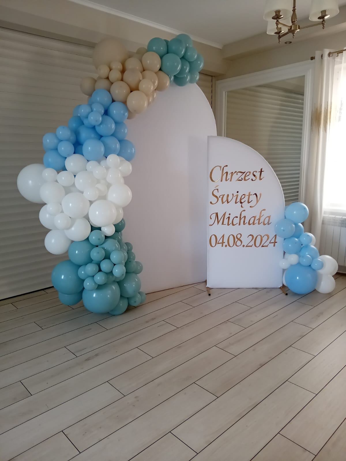 Dekoracja balonowa z białym panelem z napisem 'Chrzest Święty Michała 04.08.2024' na drewnianej podłodze.