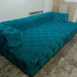 Turkusowa, pikowana sofa typu Chesterfield w warsztacie, widoczne ślady pracy na podłodze i narzędzia w tle.