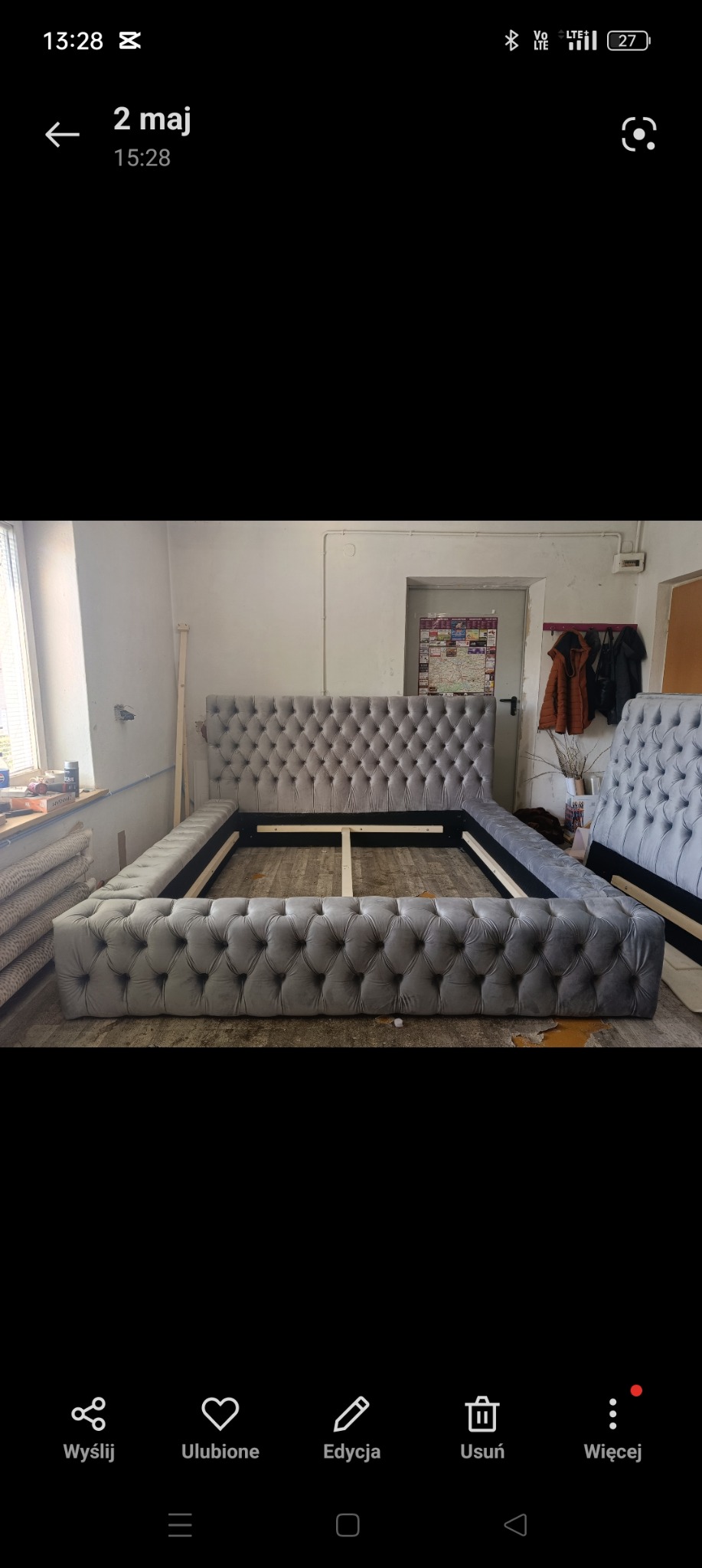 Szare, tapicerowane łóżko z pikowanym zagłówkiem w trakcie montażu, widoczne elementy konstrukcyjne ramy i drewniane listwy, w tle ściana warsztatu z narzędziami i ubraniami.