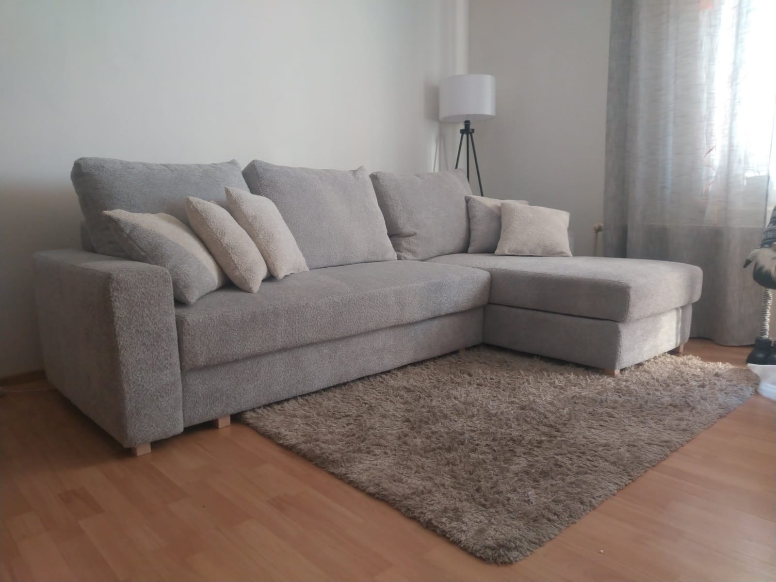 Jasnoszara, narożna sofa z miękkiej tkaniny, z puszystym dywanem na drewnianej podłodze, oświetlona lampą podłogową w tle.