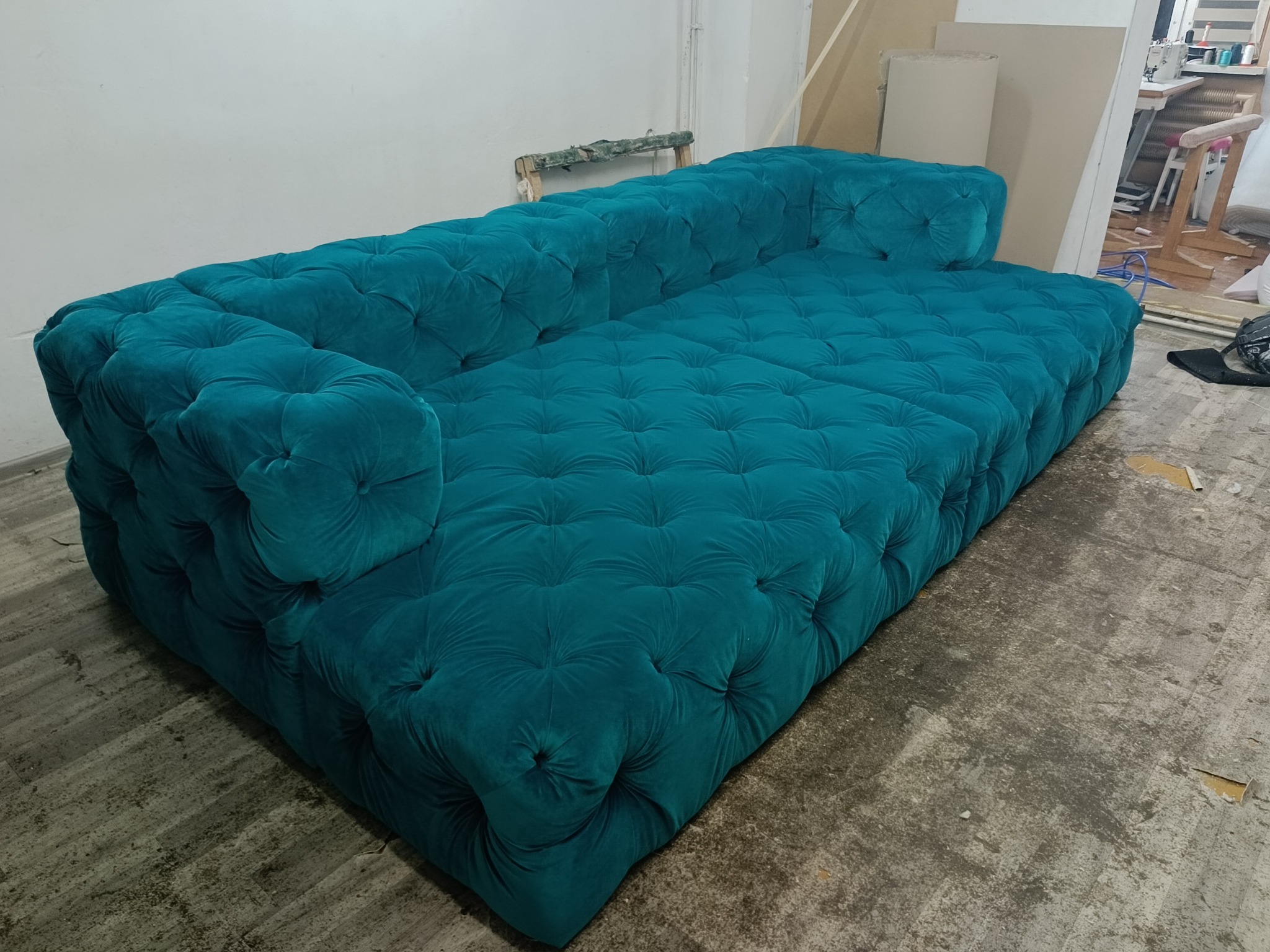 Turkusowa, pikowana sofa typu Chesterfield w warsztacie, widoczne ślady pracy na podłodze i narzędzia w tle.