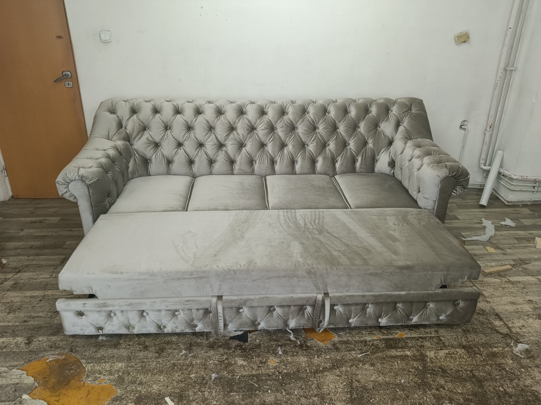 Rozłożona szara sofa Chesterfield z funkcją spania, widoczna w pomieszczeniu w trakcie renowacji w Barwałdzie Średnim, z wyraźnymi śladami użytkowania na podłodze.
