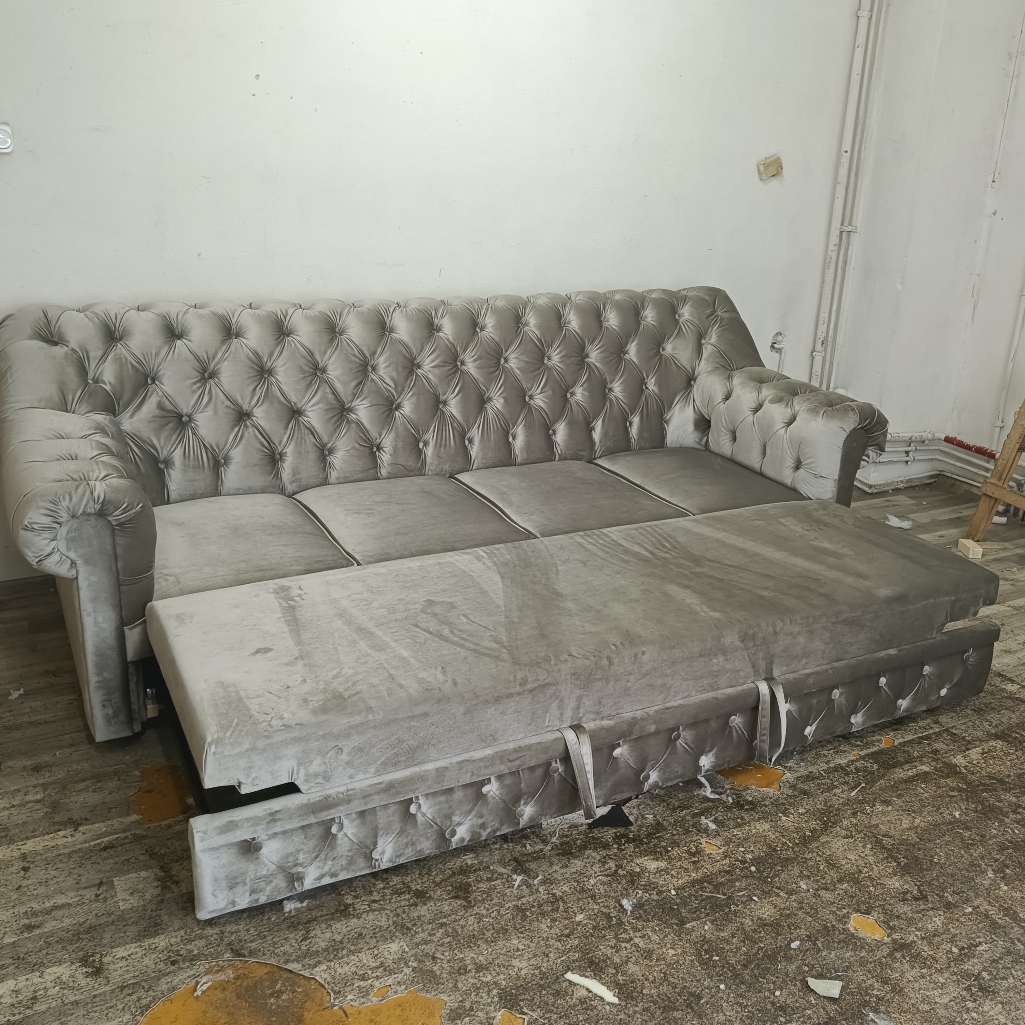 Rozłożona, szara, tapicerowana sofa z pikowaniem chesterfield na tle białej ściany i zniszczonej podłogi.