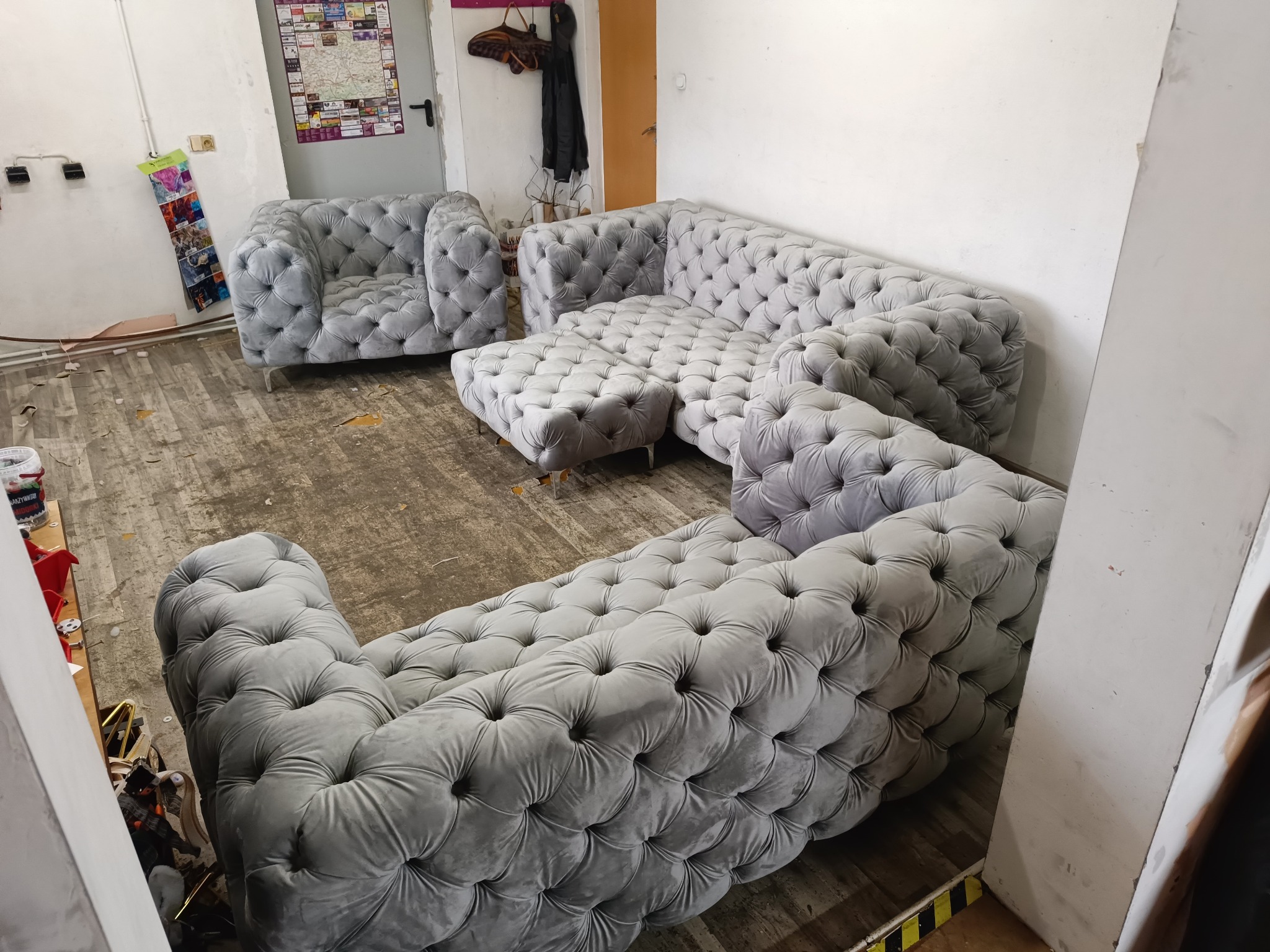 Komplet wypoczynkowy w stylu chesterfield, obity szarym welurem, składający się z narożnika, pufy i fotela, sfotografowany w pomieszczeniu w trakcie lub tuż po renowacji. Widoczne ślady prac...