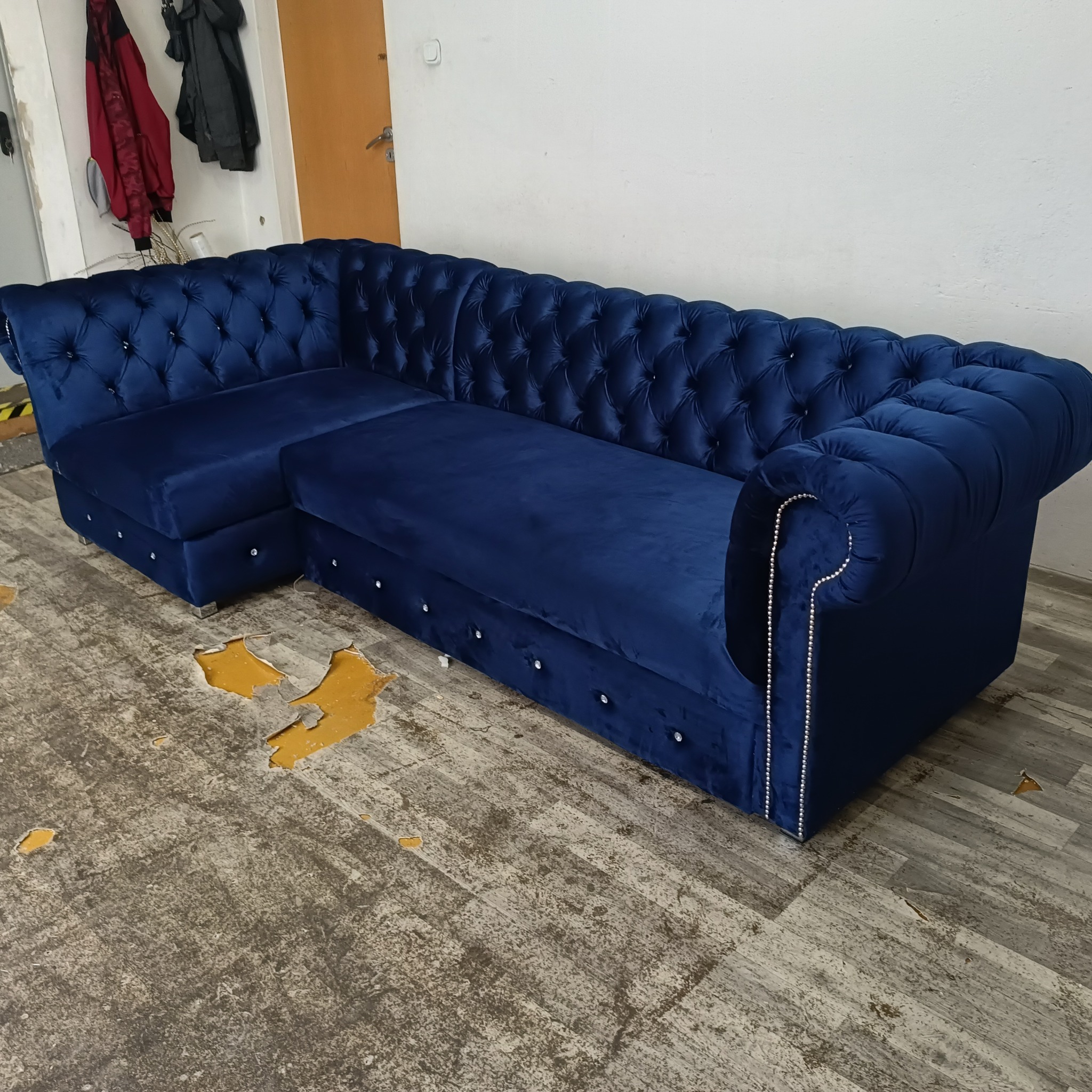 Narożnik tapicerowany aksamitem w odcieniu głębokiego granatu, z pikowaniem typu chesterfield i srebrnymi pinezkami na krawędziach, ustawiony na podłodze z widocznymi uszkodzeniami.