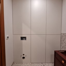 MasrockMeble - Biała zabudowa meblowa w łazience, widoczne otwory na instalacje i fragment szafki pod umywalkę w kolorze ciemnego drewna z okrągłymi, złotymi uchwytami.