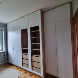 MasrockMeble - Biała szafa wnękowa z drewnianymi elementami wykończeniowymi, widoczne półki i szuflady, parkiet na podłodze częściowo zabezpieczony kartonem.