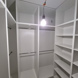 MasrockMeble - Wnętrze garderoby z białymi półkami i drążkami na ubrania, oświetlone wiszącą żarówką, widoczne kable elektryczne, marmurowa podłoga.