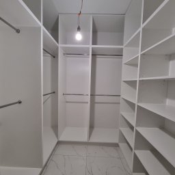 MasrockMeble - Wnętrze białej, nowoczesnej garderoby na wymiar w Ostrołęce z półkami i drążkami, oświetlonej pojedynczą żarówką zwisającą z sufitu, widok z perspektywy osoby stojącej w środku.