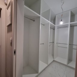 MasrockMeble - Wnętrze garderoby z białymi półkami i drążkami na ubrania, widoczne marmurowe płytki podłogowe i surowe oświetlenie z żarówką na kablu.