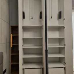 Wnętrze dwudrzwiowej, białej szafy z półkami i szufladami, widoczne drążki do wieszania ubrań, obok regał z drewnianymi półkami.