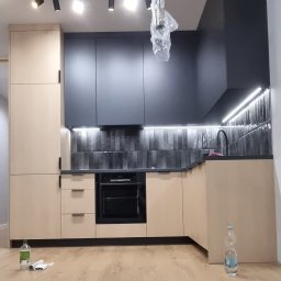 MasrockMeble - Nowoczesna kuchnia w trakcie montażu: beżowe szafki dolne, grafitowe szafki górne, podświetlenie LED nad blatem, podłoga z paneli, lampa sufitowa w folii ochronnej, widoczny kabel zasilający...