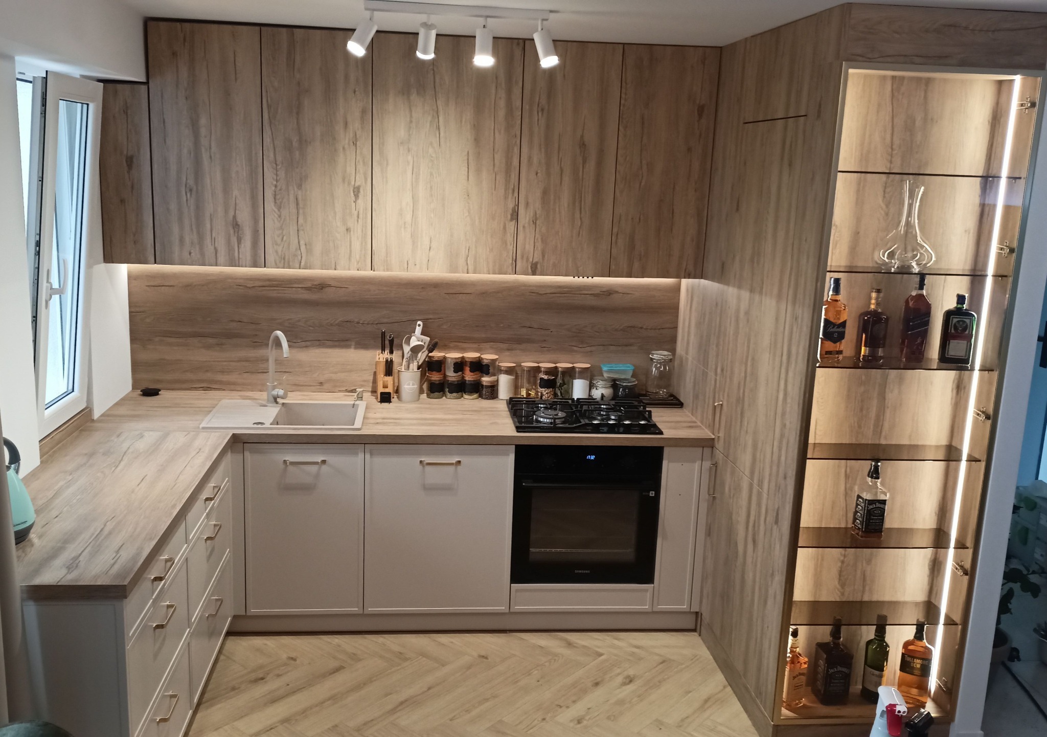 Nowoczesna kuchnia z drewnianymi frontami i blatem, jasnymi szafkami dolnymi, oświetleniem punktowym i przeszkloną witryną z alkoholem.