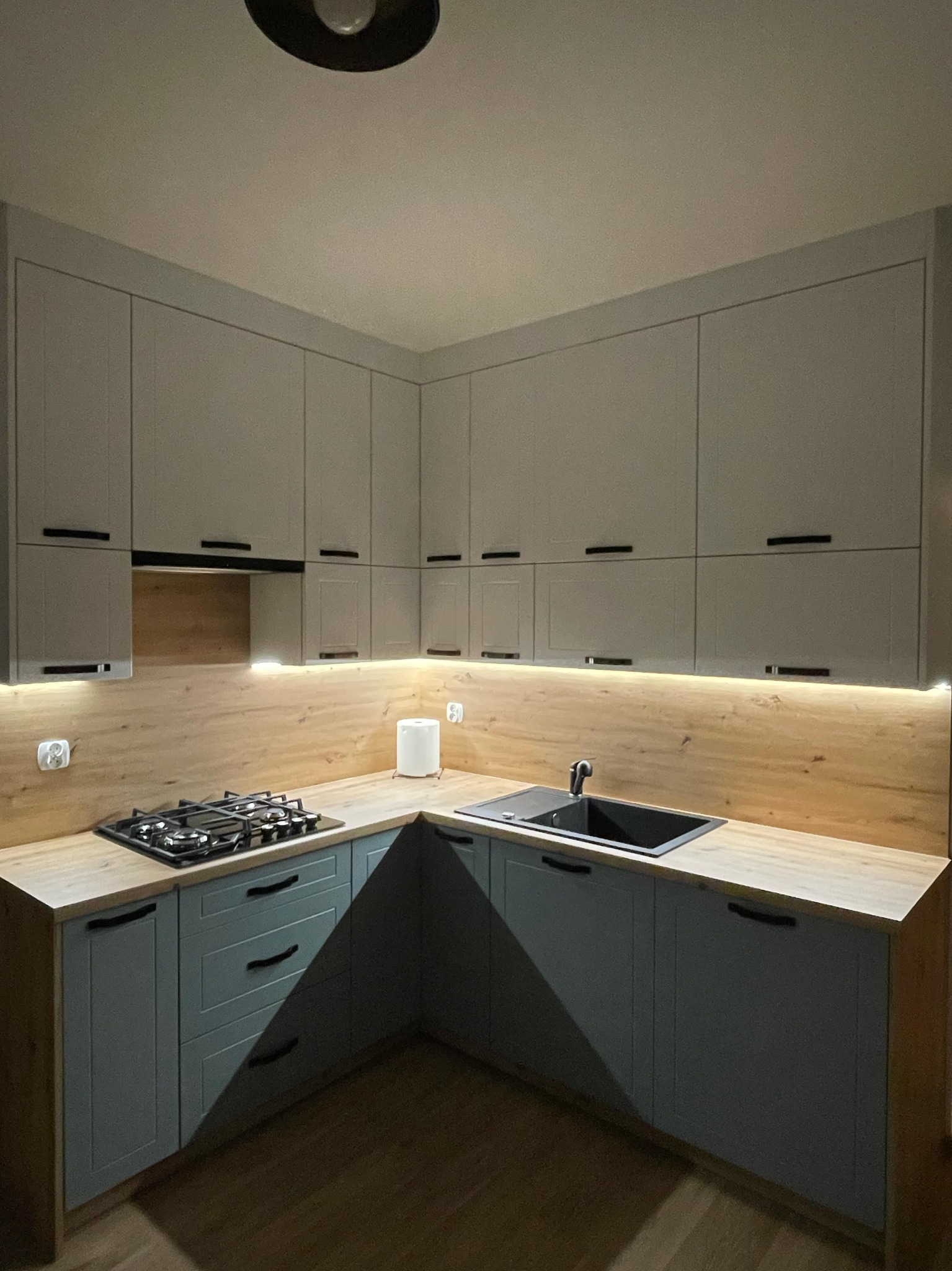 Kąt ujęcia na szaro-niebieską kuchnię z drewnianym blatem, oświetleniem LED pod szafkami i czarnym zlewem oraz płytą gazową.