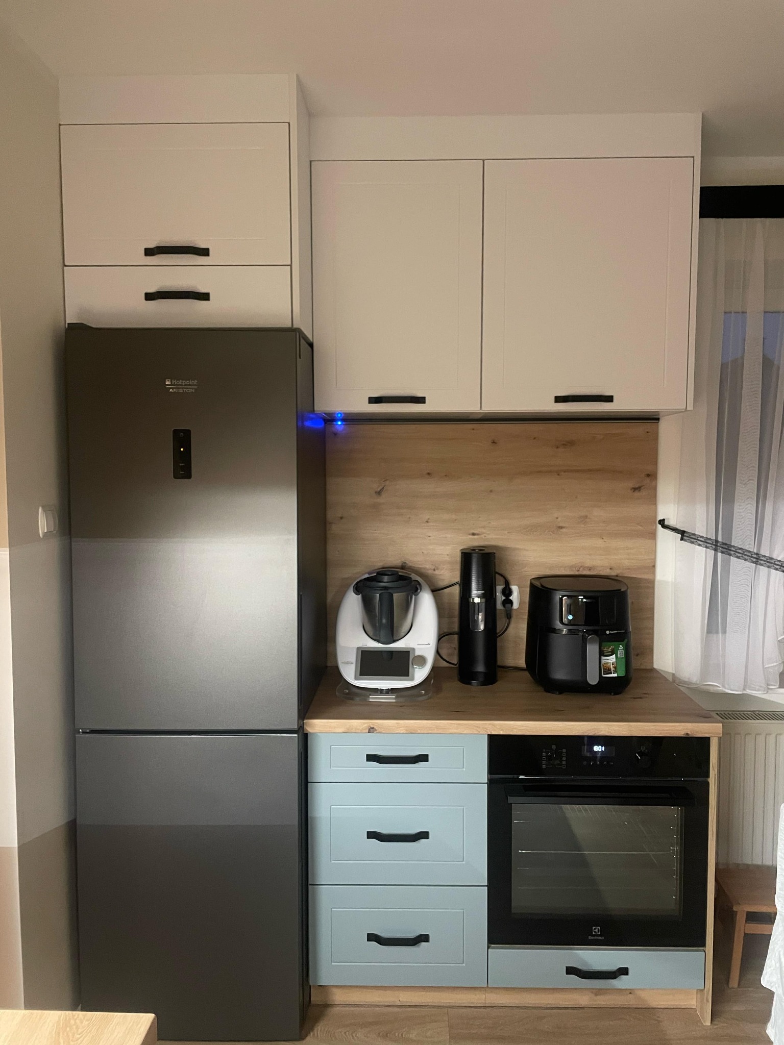 Nowoczesna kuchnia z zabudową: białe szafki górne, lodówka Hotpoint Ariston, błękitne szafki dolne z piekarnikiem Electrolux, drewniany blat i sprzęty AGD na blacie.