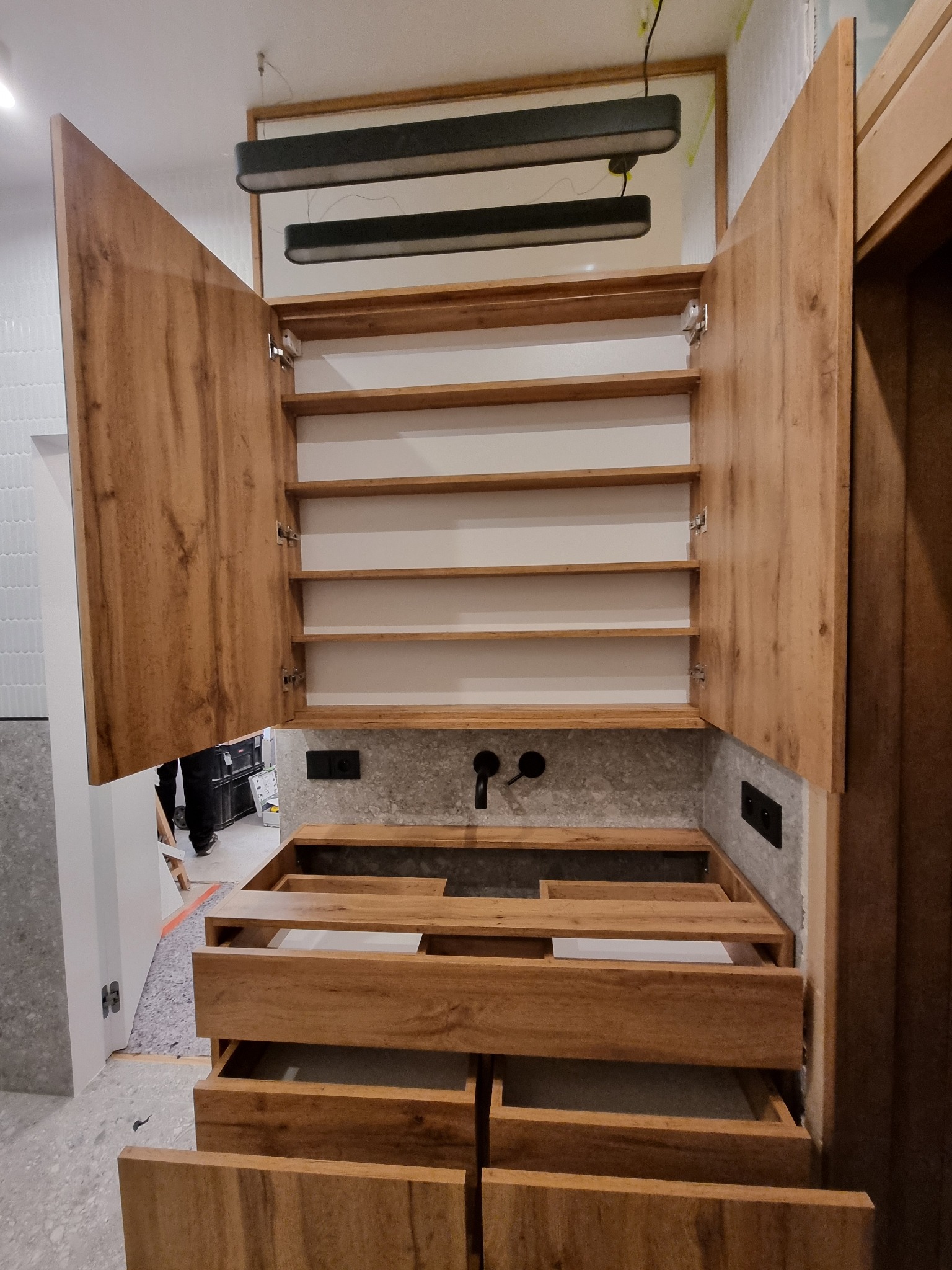 Montaż szafki kuchennej z drewnianymi frontami i półkami, widoczne otwarte drzwi szafki, niezamontowana umywalka i niedokończone szuflady, w tle białe płytki na ścianie.