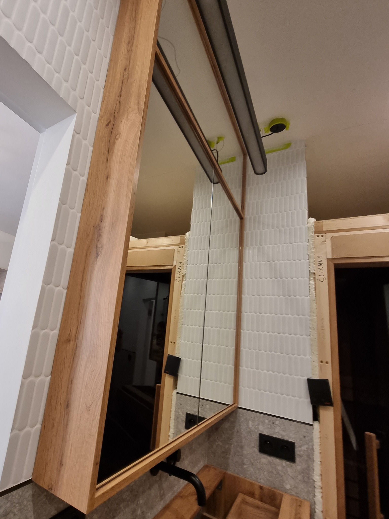 Montaż szafki z lustrem w łazience, widoczne płytki w kształcie łezki, drewniane elementy konstrukcyjne i oświetlenie sufitowe.