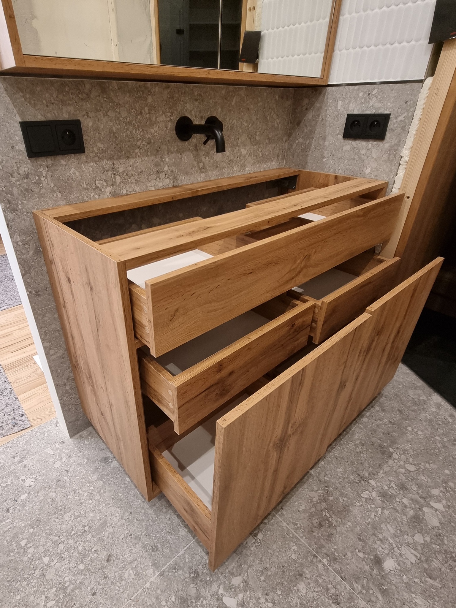 Drewniana szafka łazienkowa z wysuwanymi szufladami, zamontowana pod umywalką, z widoczną instalacją wodną i elektryczną w tle.