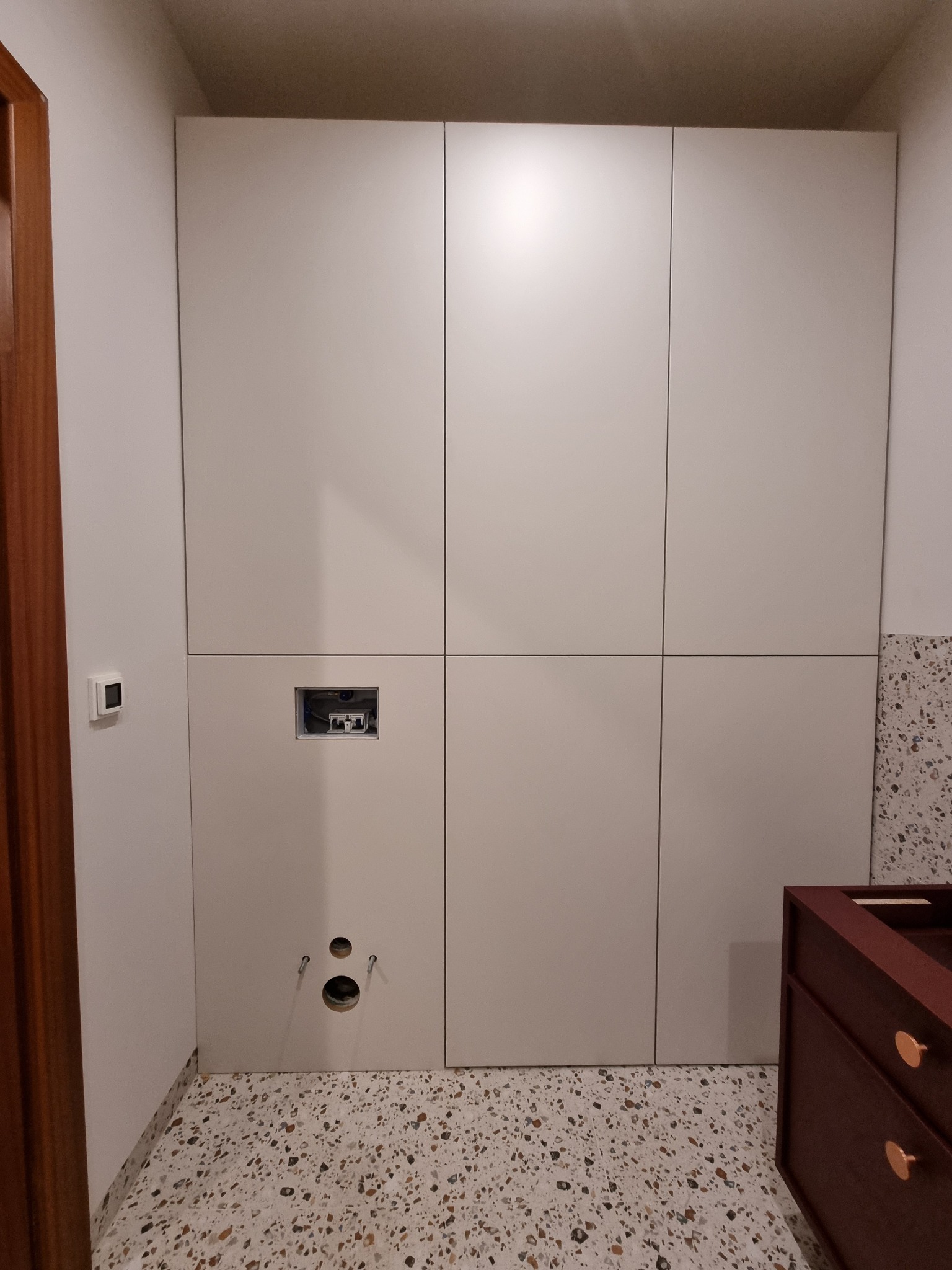 Biała zabudowa meblowa w łazience, widoczne otwory na instalacje i fragment szafki pod umywalkę w kolorze ciemnego drewna z okrągłymi, złotymi uchwytami.