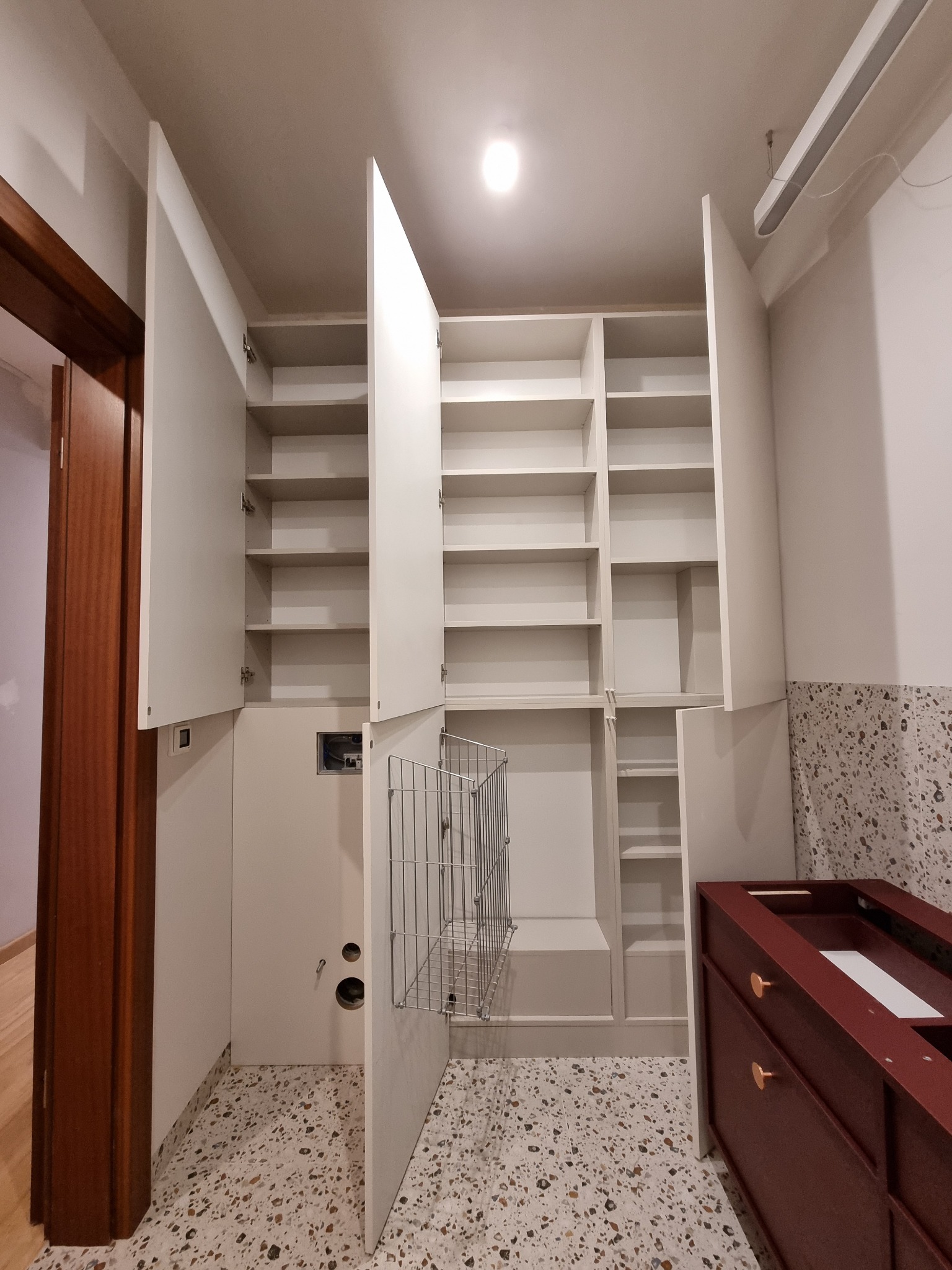 Wysoka, biała szafa wnękowa z otwartymi drzwiami ukazującymi puste półki i metalowy kosz na pranie, z widocznym fragmentem ciemnoczerwonej szafki z szufladami w pomieszczeniu z podłogą lastryko.
