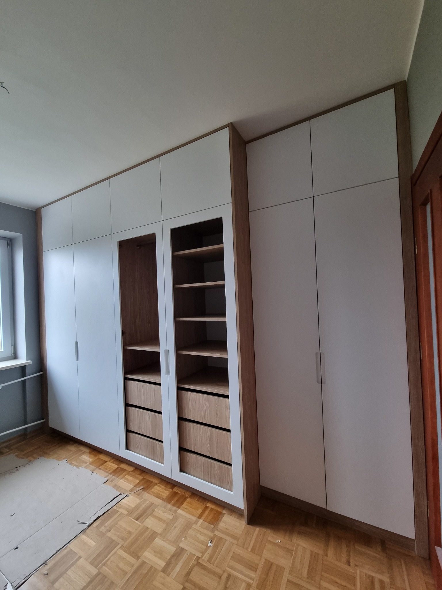 Biała szafa wnękowa z drewnianymi elementami wykończeniowymi, widoczne półki i szuflady, parkiet na podłodze częściowo zabezpieczony kartonem.