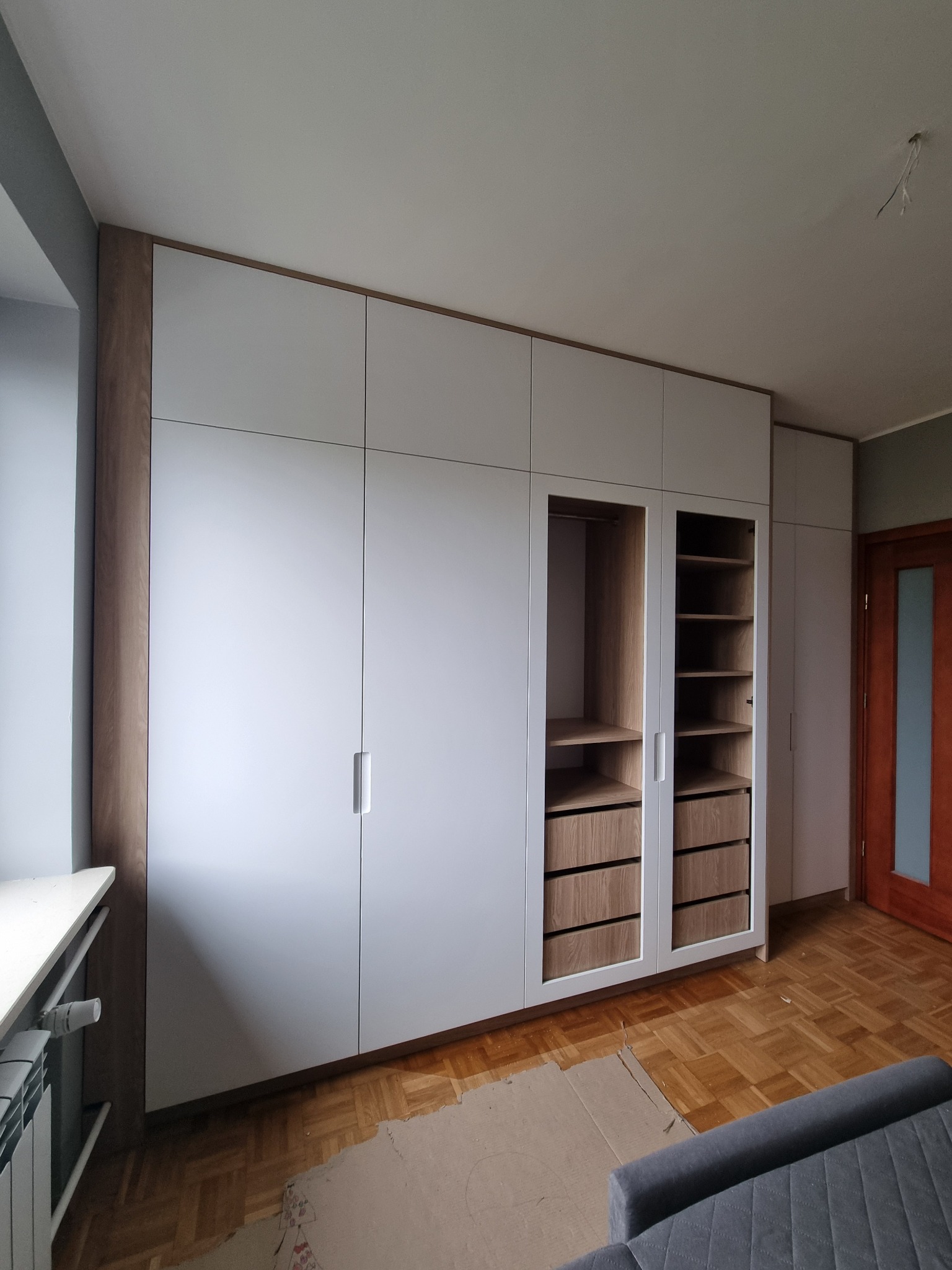 Duża, biała szafa wnękowa z drewnianymi akcentami, widoczne półki i szuflady, parkiet na podłodze, fragment sofy w kadrze.