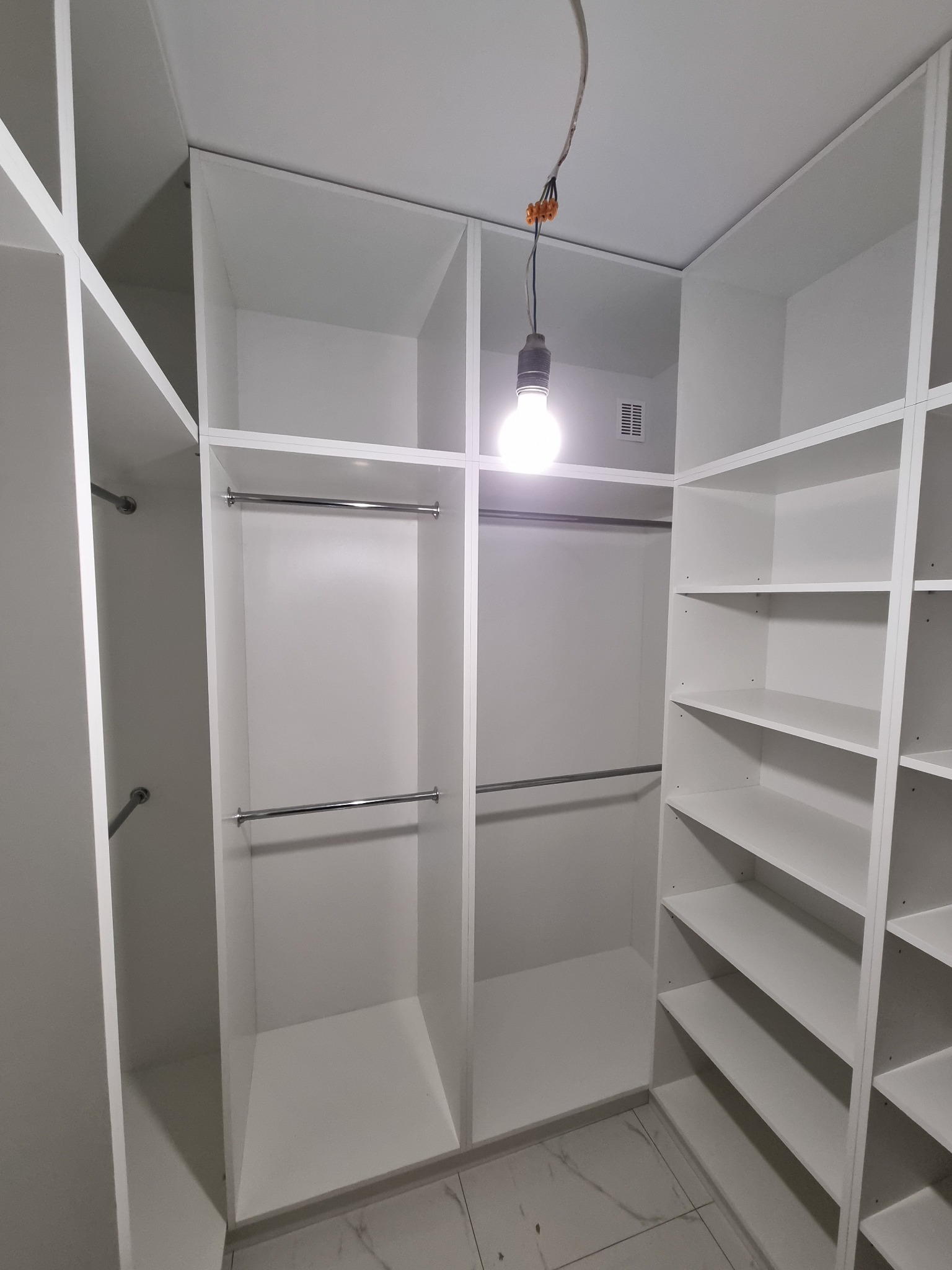Wnętrze garderoby z białymi półkami i drążkami na ubrania, oświetlone wiszącą żarówką, widoczne kable elektryczne, marmurowa podłoga.