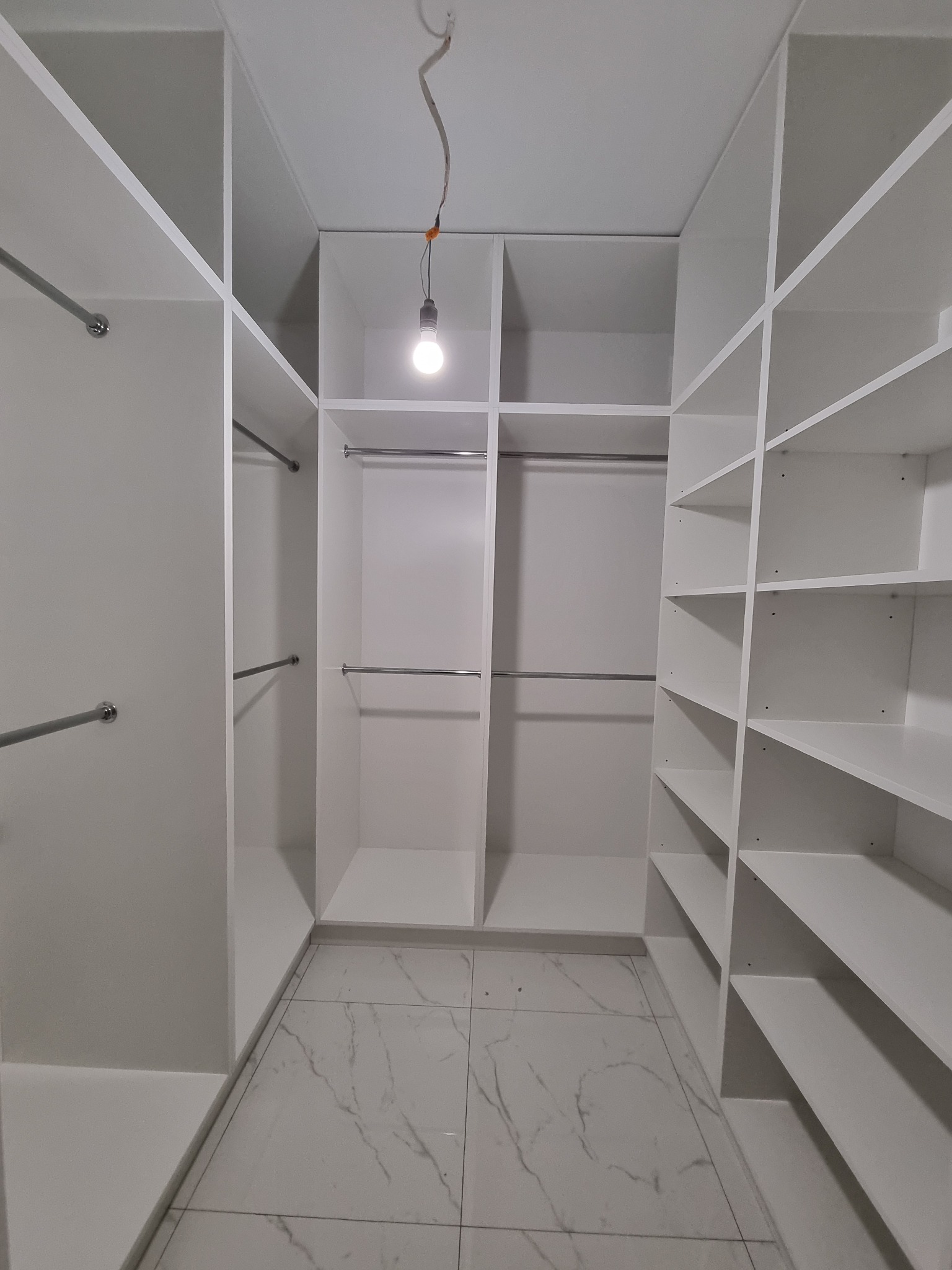 Wnętrze białej, nowoczesnej garderoby na wymiar w Ostrołęce z półkami i drążkami, oświetlonej pojedynczą żarówką zwisającą z sufitu, widok z perspektywy osoby stojącej w środku.
