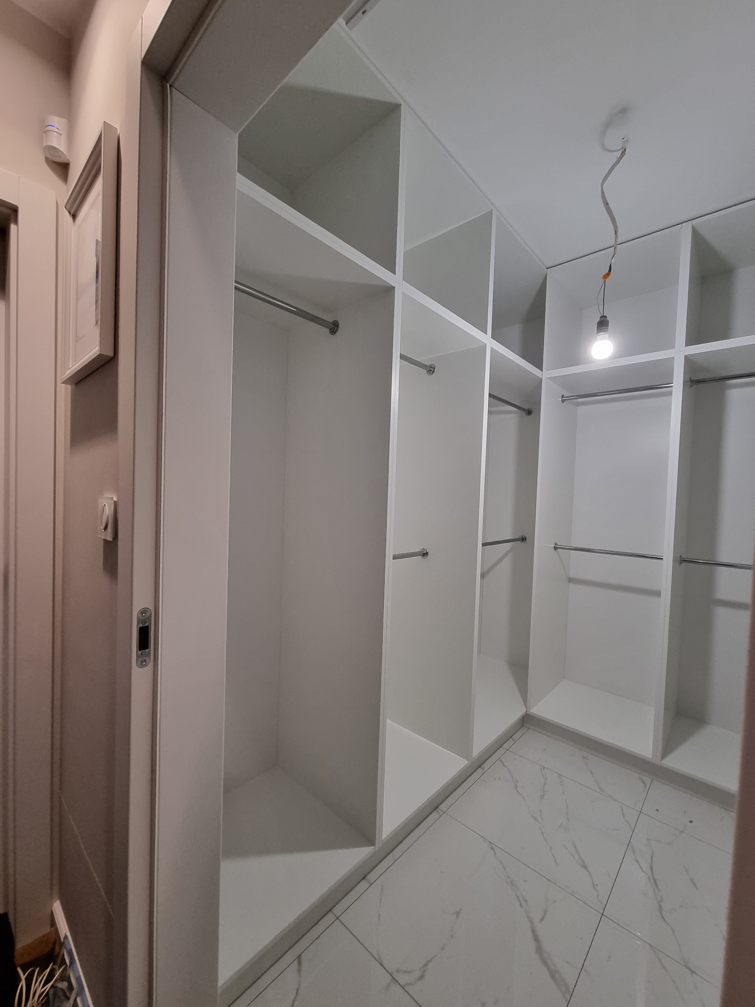 Wnętrze garderoby z białymi półkami i drążkami na ubrania, widoczne marmurowe płytki podłogowe i surowe oświetlenie z żarówką na kablu.