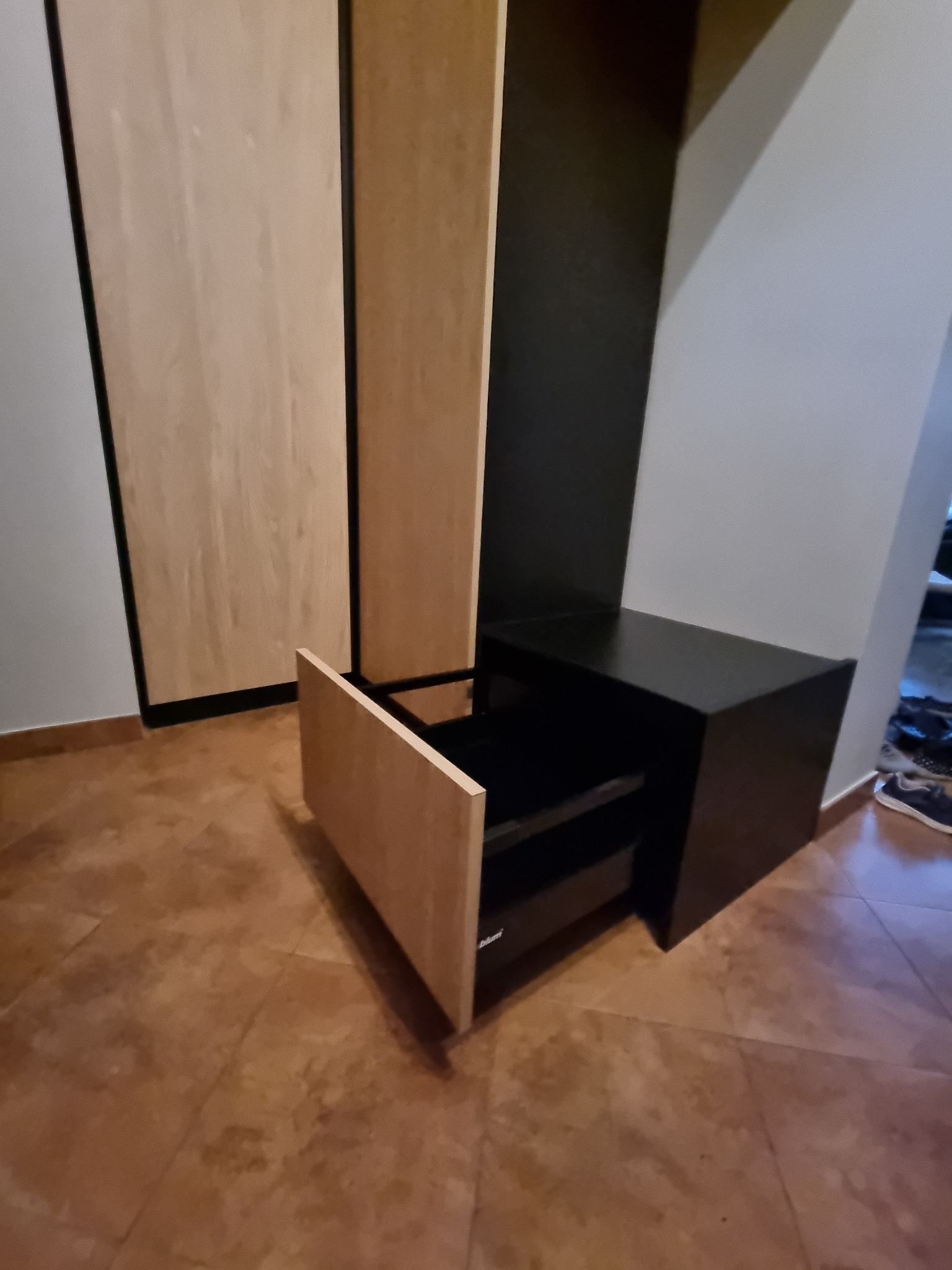 Fragment szafy z jasnymi frontami i czarnym korpusem, szuflada otwarta, widoczny system Blum, na brązowej podłodze.