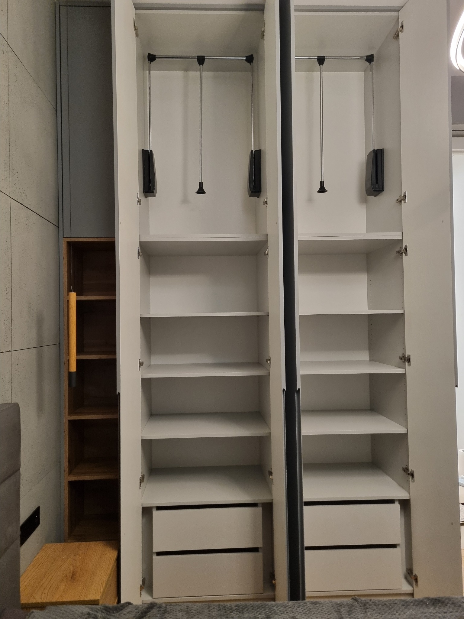 Wnętrze dwudrzwiowej, białej szafy z półkami i szufladami, widoczne drążki do wieszania ubrań, obok regał z drewnianymi półkami.