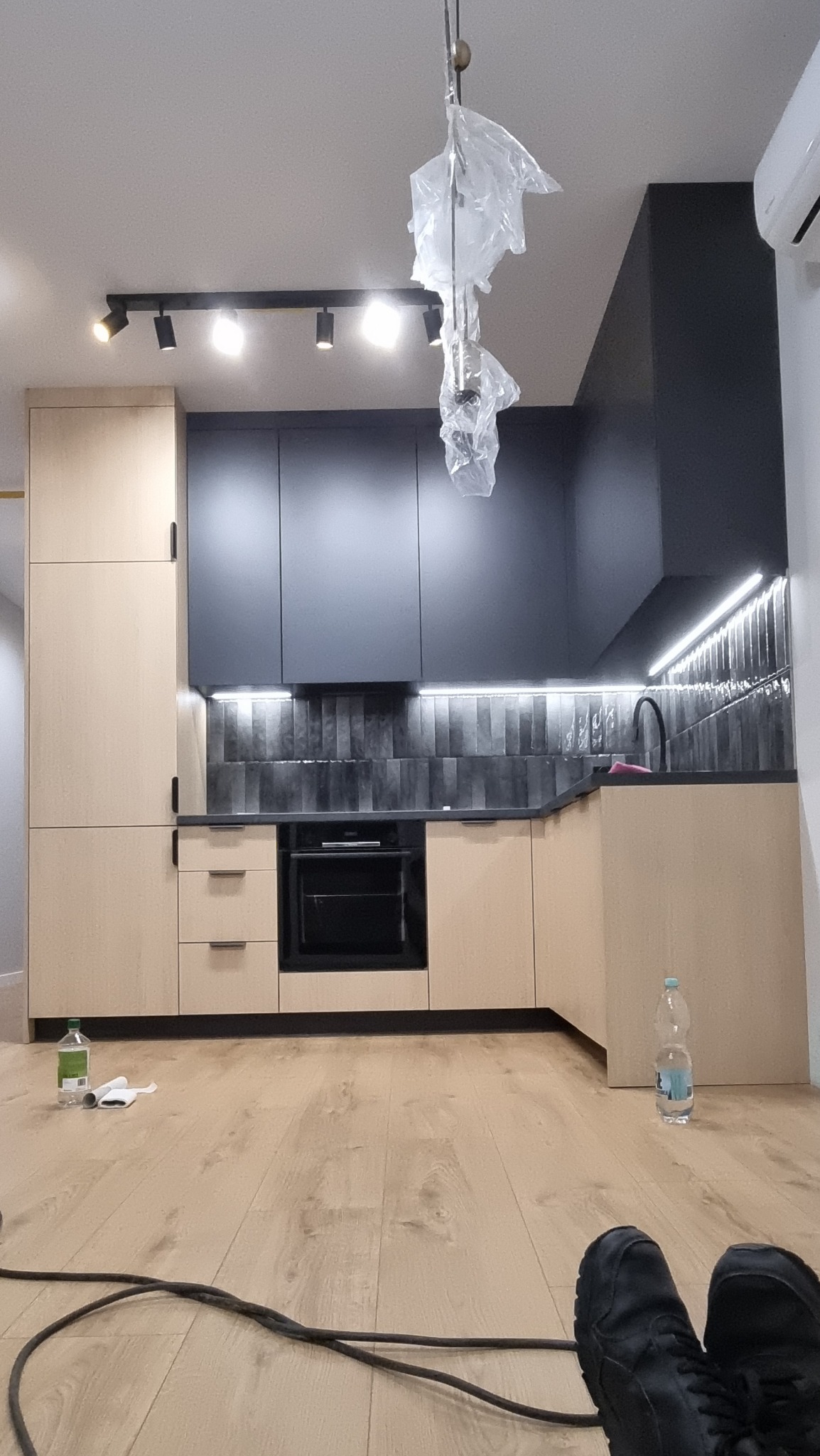 Nowoczesna kuchnia w trakcie montażu: beżowe szafki dolne, grafitowe szafki górne, podświetlenie LED nad blatem, podłoga z paneli, lampa sufitowa w folii ochronnej, widoczny kabel zasilający...
