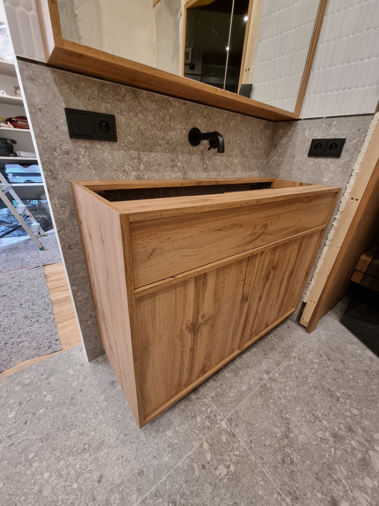 Drewniana szafka umywalkowa z szufladą i blatem, czarna bateria ścienna, szara ściana z gniazdkami, w tle drabina i płytki w kształcie rybiej łuski, stan surowy.