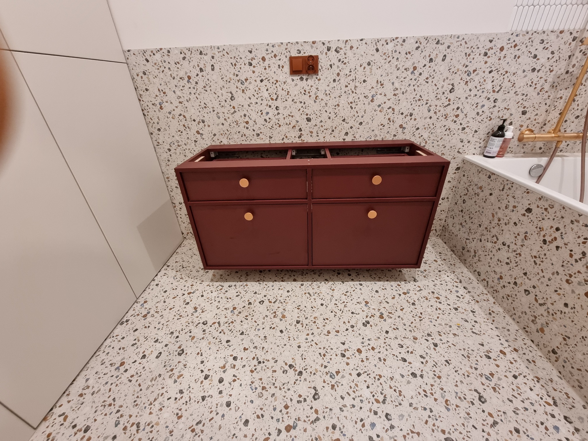 Czerwona szafka pod umywalkę z czterema szufladami i złotymi uchwytami, zamontowana na ścianie z wzorem lastryko, widok z dołu.